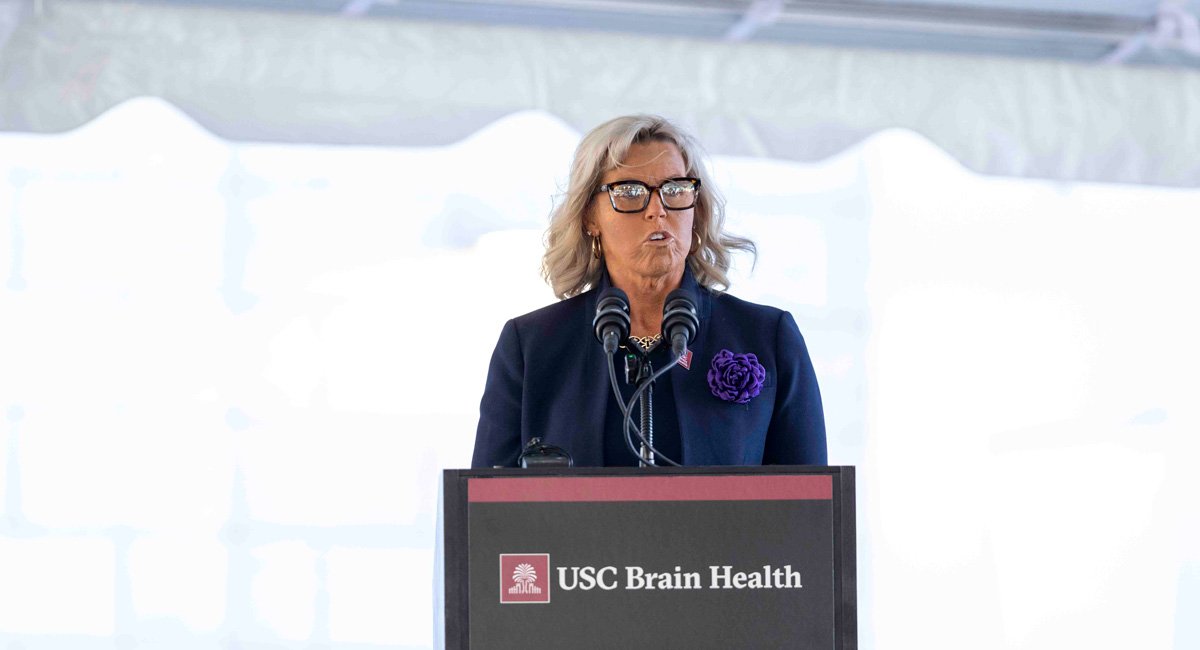 Laura Haynes, a woman in a purple blouse, stands behind the podium at the USC Brain Health Center giving a speech.