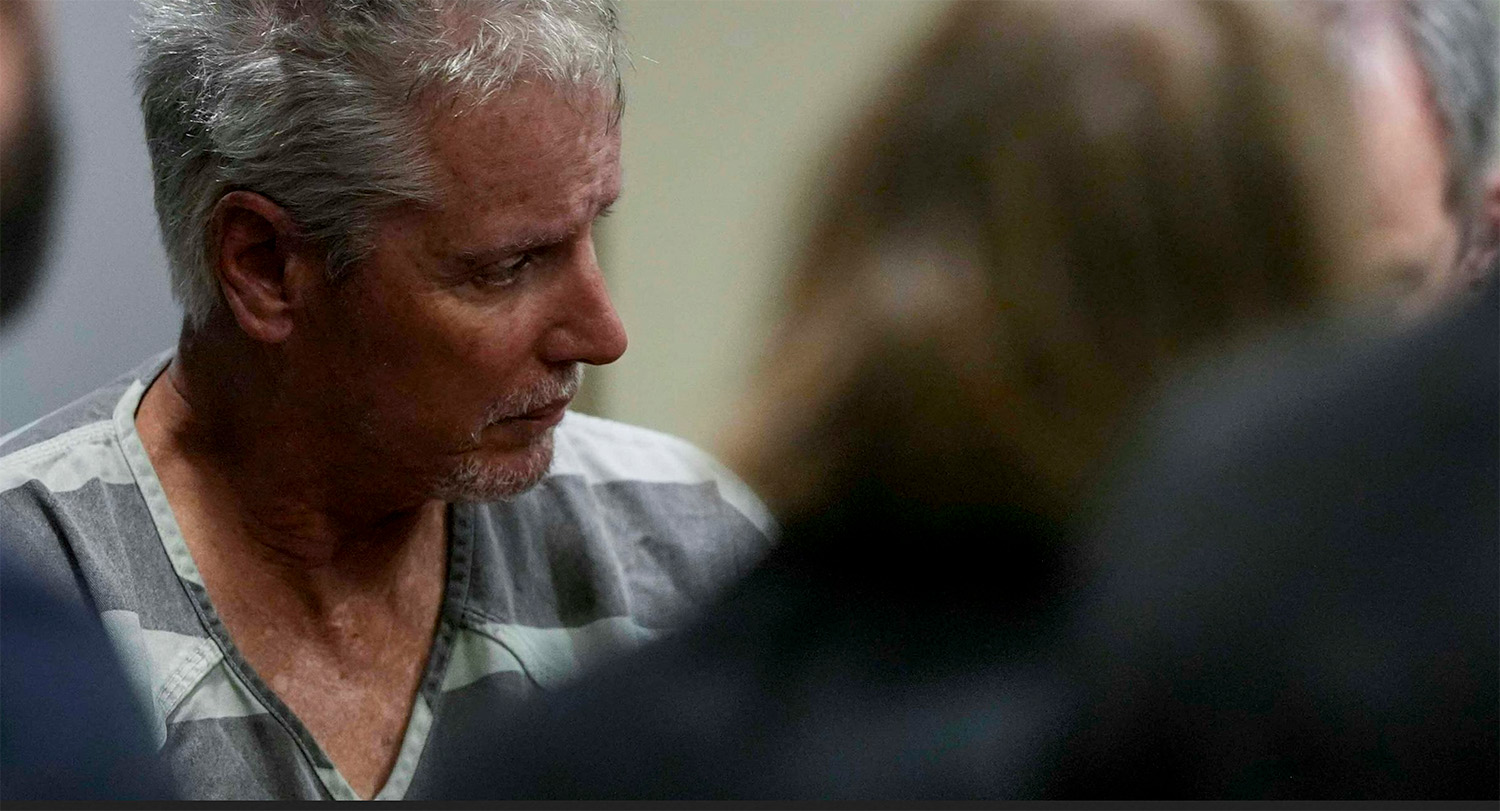 A gray-haired white male in striped prison garb surrounded by people in suits in courtroom setting