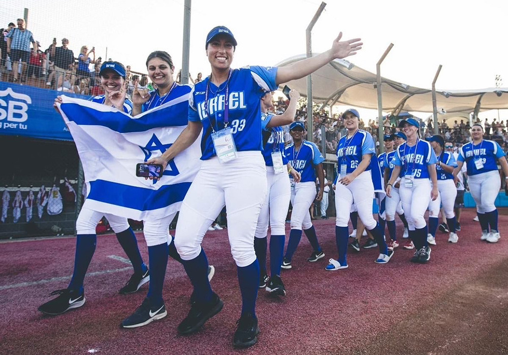 Israel softball players celebrate on the field, holding a national flag during an international competition.