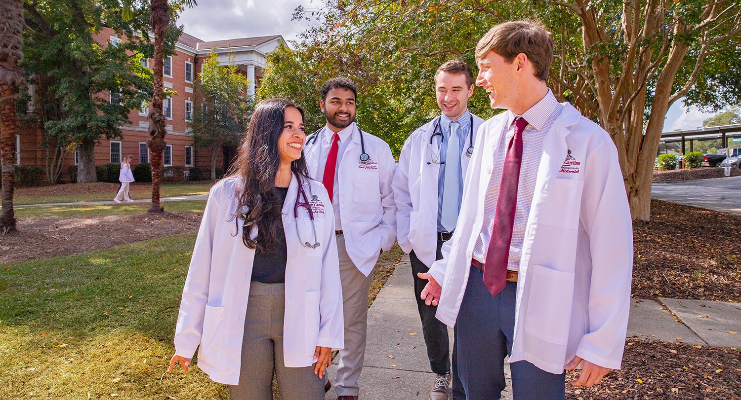 Four medical students in white lab coats walk and talk on a campus pathway lined with trees and academic buildings.