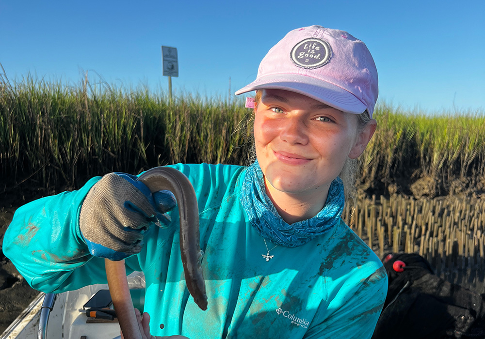 Young woman in blue shirt and ponytail holding a snake in a coastal marsh setting
