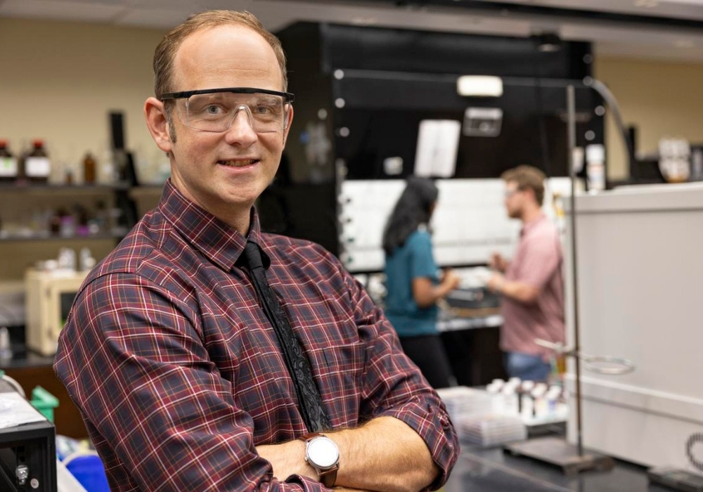 Morgan Stefik smiles in his chemistry lab, where two graduate students discuss in the background.