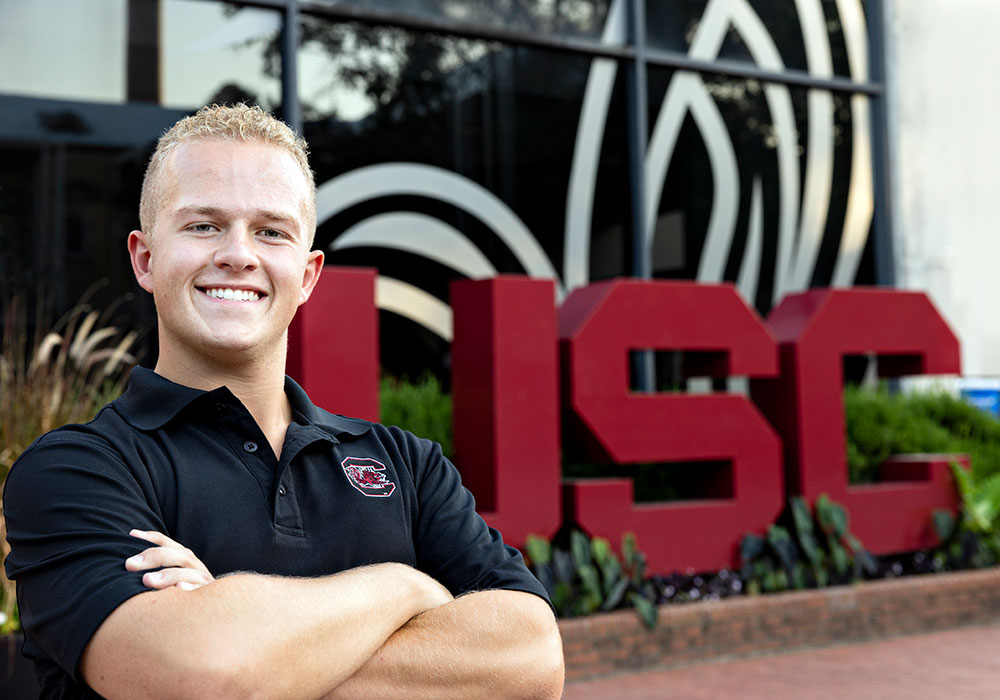 A smiling man in USC polo shirt poses with crossed arms in front of university signage.