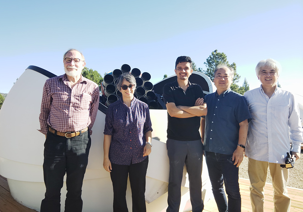 A group of scientists stand in front of a large telescope on a bright day.