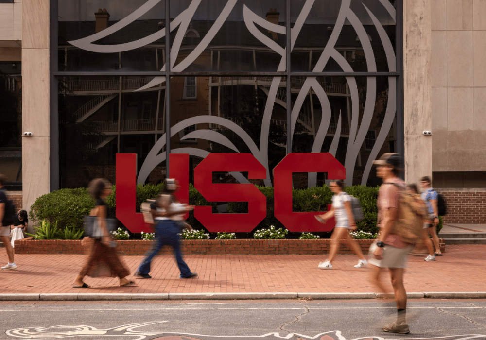 University of South Carolina (USC) students walk past a large USC logo outside a campus building. A graphic reads, “U.S. News Nation’s Best First-Year Experience — Public Universities.”