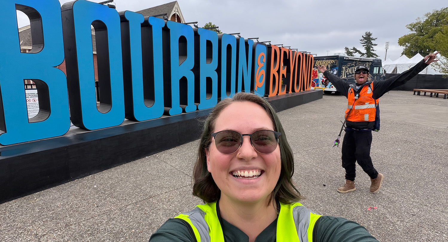 Woman in sunglasses and yellow utility vest poses before 'Bourbon and Beyond' festival sign with playful coworker gesturing in background.
