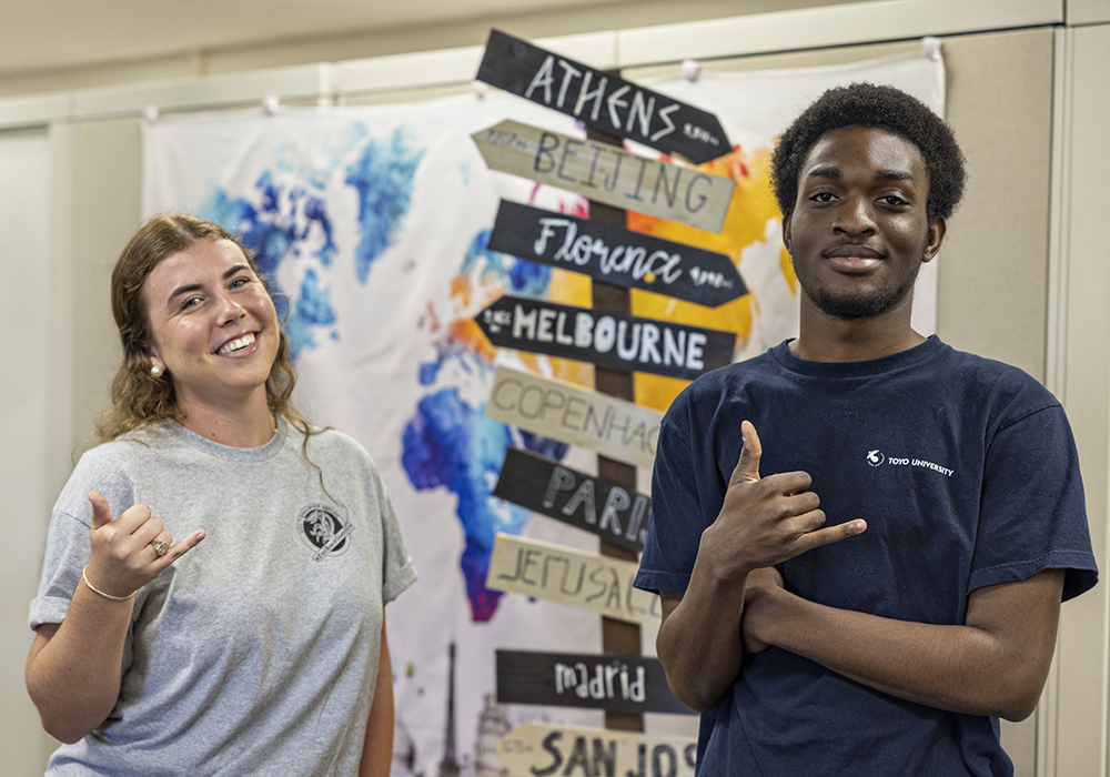 Kelcie Pollinger and Isaiah Smalls pose with 'spurs up' hand signals in front of an artistic sign pointing to multiple place names: Athens, Beijing, Florence, Melbourne, Copenhagen, Paris, Jerusalem, Madrid, San Jose...