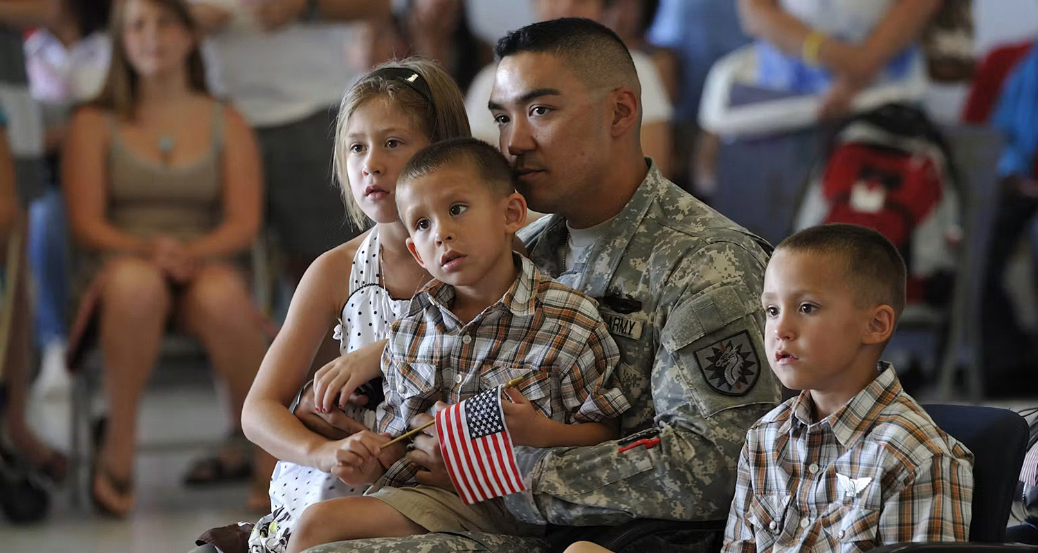 A man wearing a military uniform sits and holds a small boy on his lap, while a girl and boy sit close to him.