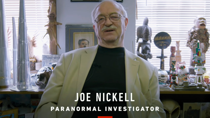 a man sits in front of a desk full of weird objects; screen title reads: Joe Nickell, paranormal investigator