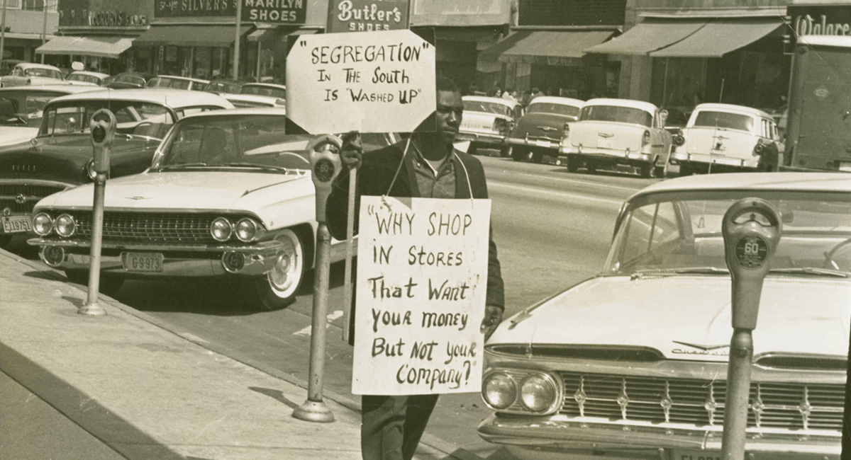 A man, possibly Gene Hawkins with 2 protest signs walking down Main Street, in which is lined with cars. The two protest signs read: Segregation in the south is washed up and Why shop in stores that want your money but not your company? Many business and signs can be seen in the background.