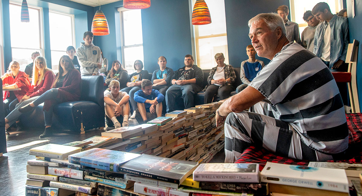 man sits on cot in a makeshift prison baracaded in by banned books 