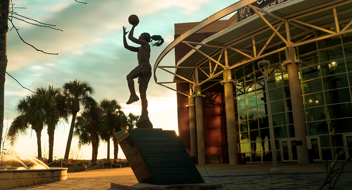 Statue of A'ja Wilson, female basketball player, mid jump shooting a basketball in front of a sunset at the Colonial Life Arena. 