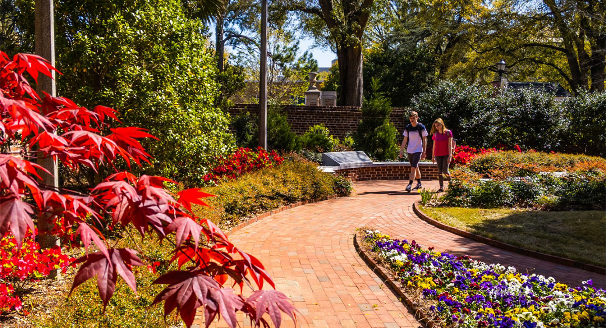 Two students walking on the brick pathway in the Desegregation Garden surronded by flowerning plands and beautiful red trees.