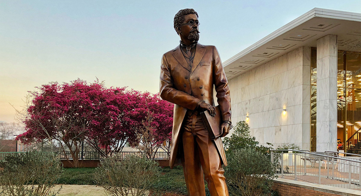 Statue of Richard T. Greener, the first African-American professor at the University of South Carolina, with pink flowers blooming and a sunset in the background. 