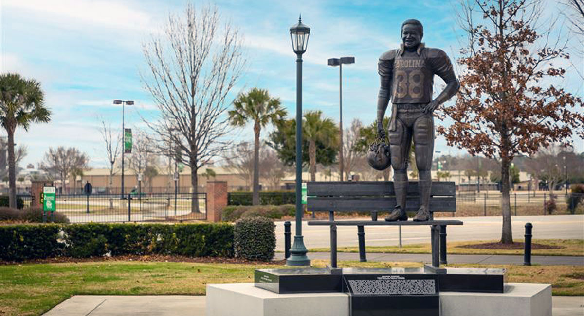 George Rogers statue fearues him in full football uniform holding a helment standing on a bench outside of Williams Brice Stadium. 