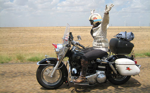 A side view of Kay Andrea raising her hands in the air while riding her vintage bike on Route 66