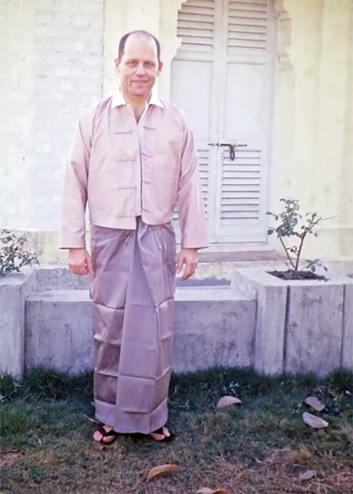 1960s-era faded photo print of a man wearing blue sari with pink shirt standing before yellow building