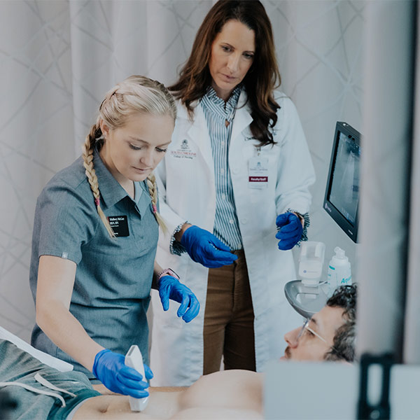  two women nurses stand at hospital bedside, one using a portable ultrasound on a patient