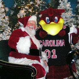 A rosy cheeked Santa sitting in front of a decorated Christmas tree with the UofSC mascot, Cocky. 