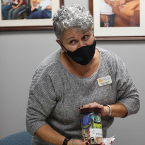 Woman with gray hair, gray shirt and black mask standing at a table, displaying health items. Man and woman at table with back to viewer.