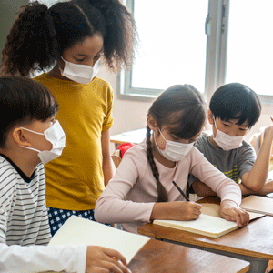 A teacher wearing a mask sits with her students at a desk