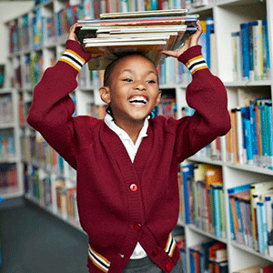 A smiling studen in a red sweater walks through a library holding a stack of books on her head.