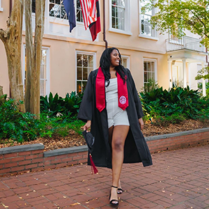 Bailey Brown, wearing a graduation robe and holding her cap, stands in front of the President's House on the historic Horseshoe