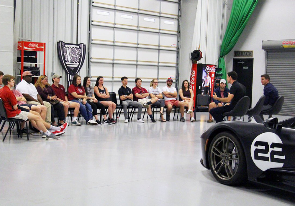 a class listening to a guest speaker by a racecar