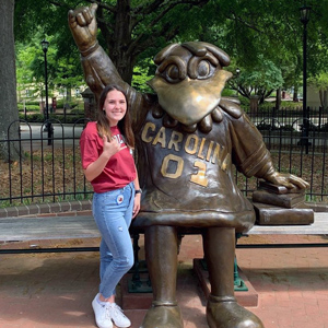 A woman in a garnet shirt poses with the Cocky statue. 