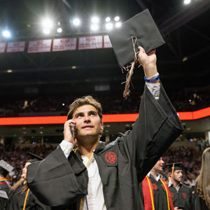 man in graduation robe holds his mortar board above his head as he talks on his phone
