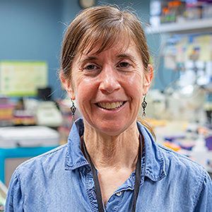 A white woman with brown hair tied back and wearing a blue button-down shirt smiles from a biology lab setting