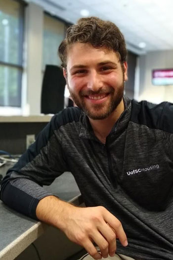Alex Kroll at a front desk of a residence hall, smiling in a uofsc housing pullover