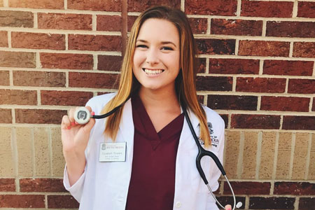 Senior Nursing major Liz Powers posing in front of a brick wall wearing her UofSC scrubs, white jacket and stethoscope