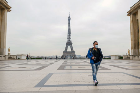 man wearing a mask walks in Paris with the Eiffel Tower in the background
