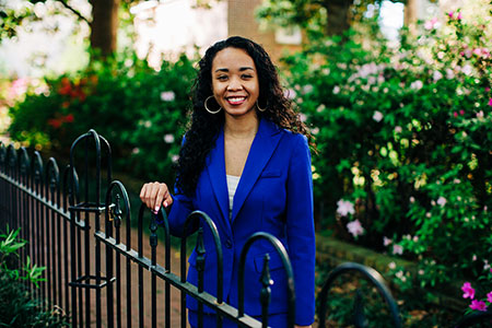 student in a blue suit standing by an iron fence with shrubbery in the background