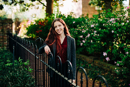 female student in a black jacket and garnet blouse standing behind an iron fence