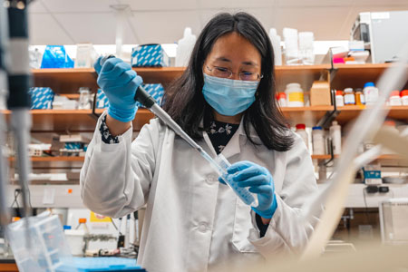 person with dark hair in a lab wearing lab coat, blue mask and blue gloves
