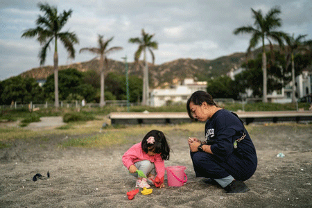 Young girl with bucket and plastic shovel plays in sand while a women watches with palm trees in the background