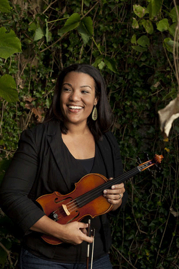 female musician smiling wearing a black dress holding a violin with vines and leaves in the background