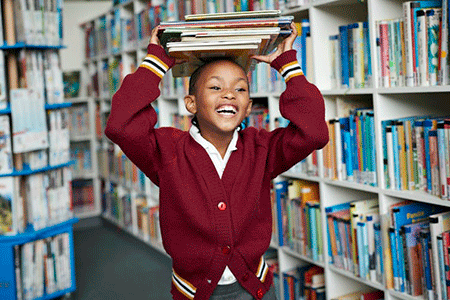 A smiling student in a red sweater walks through a library holding a stack of books on her head.