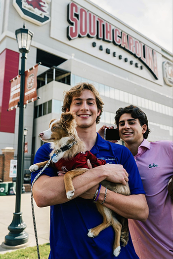 Evan Walker in front of Williams-Brice Stadium holding a brown and white dog, a man smiles behind him