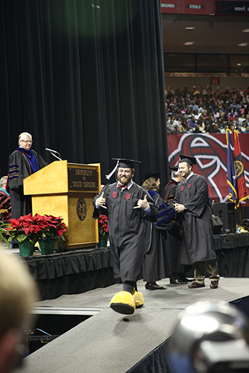 A man walks across the graduation stage with cocky mascot feet