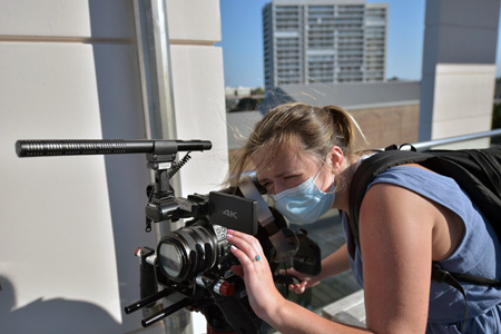 woman looks through a video camera view finder with buildings in the background