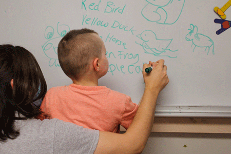 Adult in gray shirt assists student in coral shirt writing on a white board.