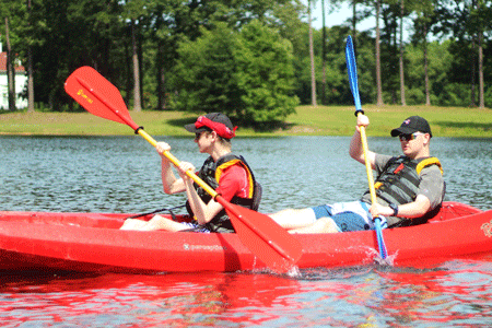 Two young men paddle a red kayak with shoreline, grass and treees in background.