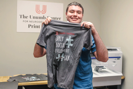Young male student in a blue shirt holds a gray T-shirt printed at The Unumb Center print shop.