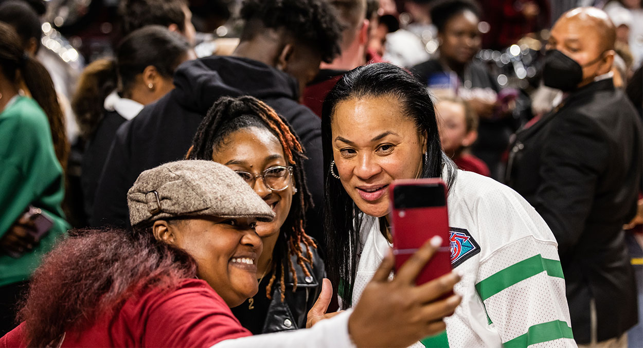 Dawn Staley taking a selfie with fans at a women's basketball game. 