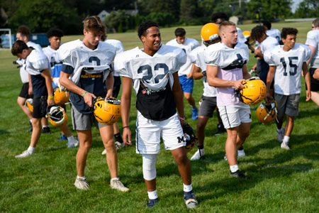 High school football players stand on field sweating and holding helmets in white uniforms