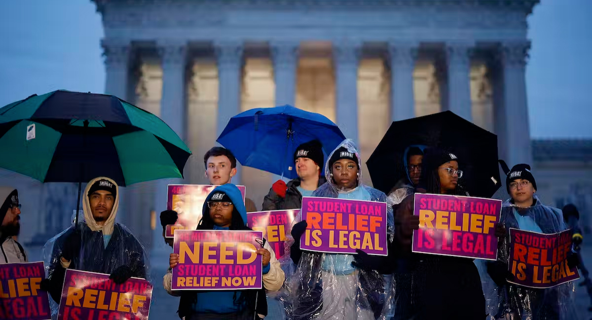 A group of nine young men and women stand in the evening rain, some hold umbrellas. Others wear hats or hoodies. Each of them holds a purple, pink, orange and yellow sign that reads: Student loan relief is legal.