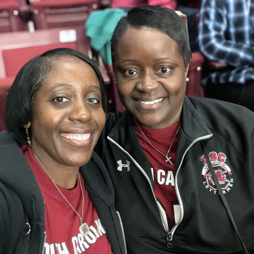 two women wearing Gamecocks clothing sit in the stands at a basketball game